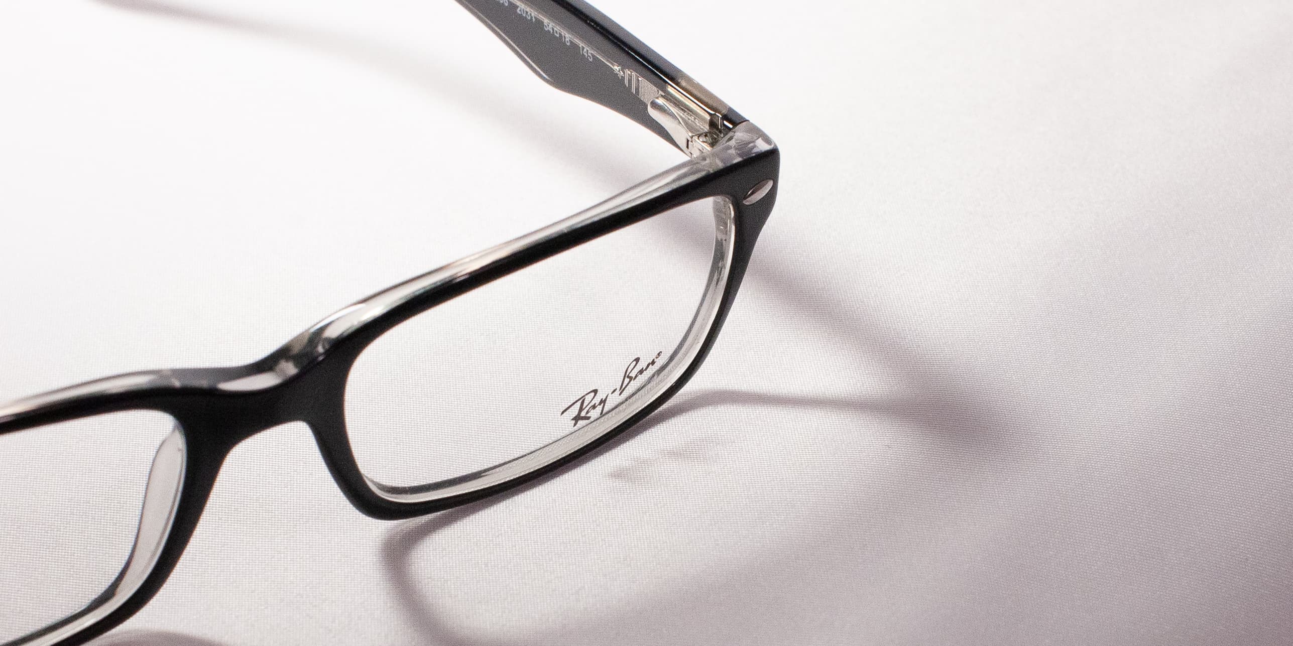 Close-up of a black plastic Ray-Ban optical frame at a low angle, soft fabric texture in the background.
