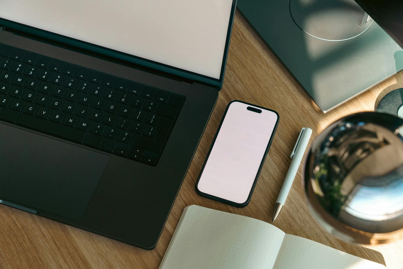Top-down workspace with a laptop, blank phone, open notebook, and pen on a wooden desk.