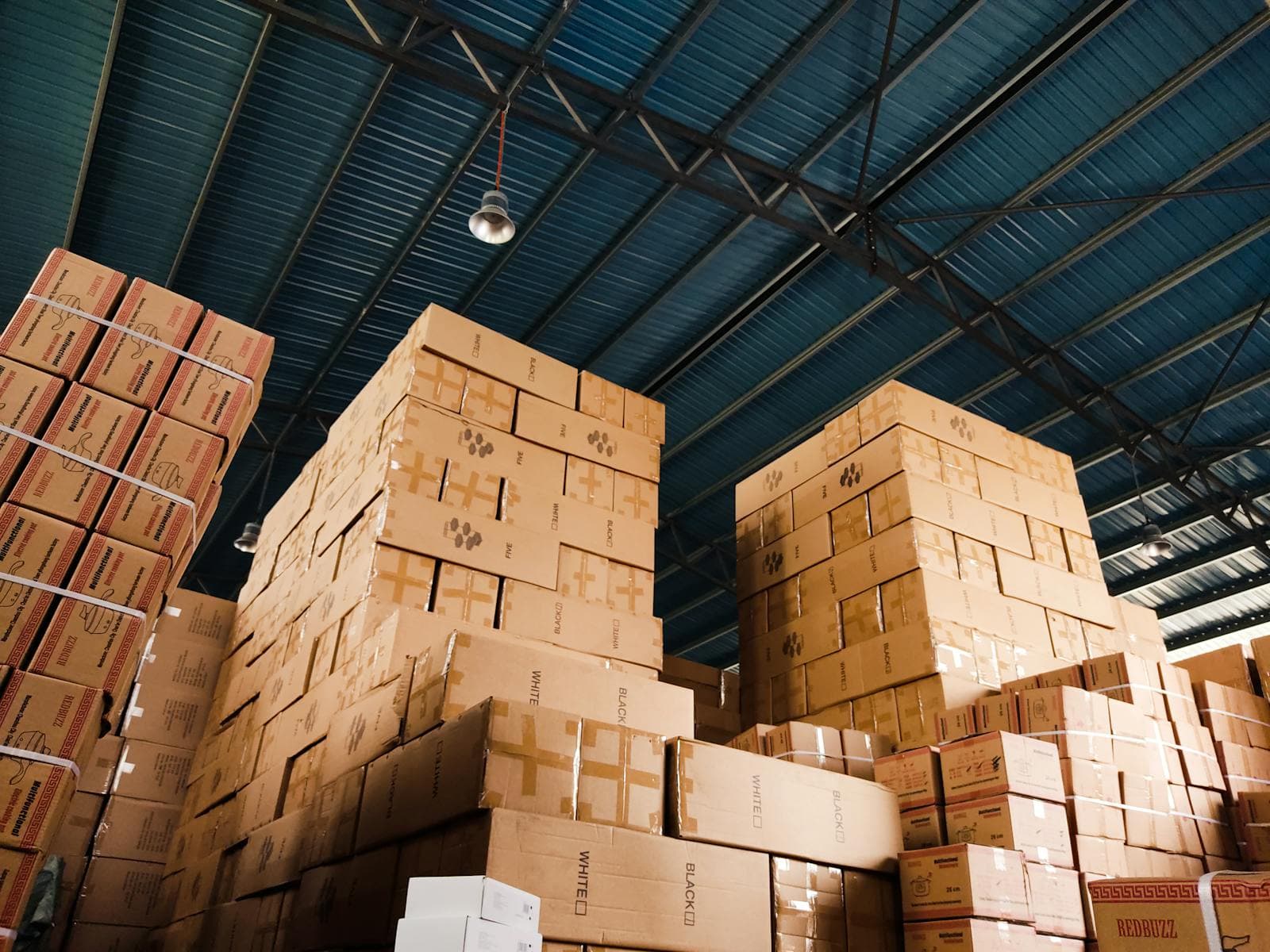 Stacks of shipping boxes inside a warehouse with a blue metal ceiling overhead.